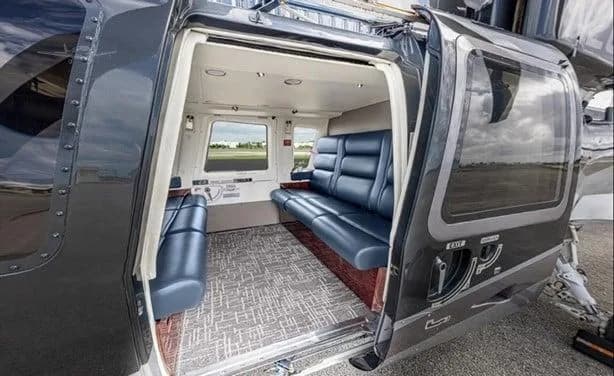 The open side door of a dark grey helicopter reveals its luxurious interior cabin with blue leather seating and a patterned carpet.