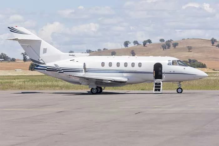 A white business jet with blue stripes on the tail and fuselage is parked on a tarmac with its main door open.