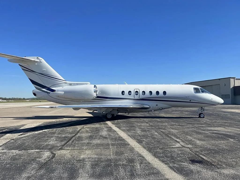 A white business jet with dark blue stripes is parked on an asphalt tarmac next to a hangar, viewed from the side.