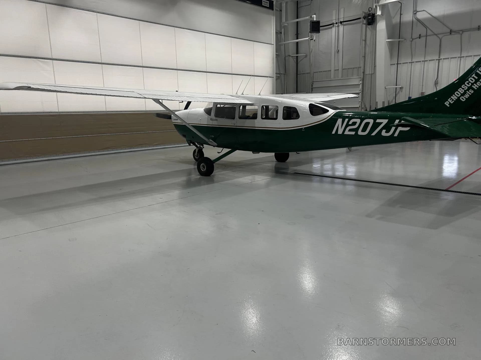 A white and green Cessna 207 Stationair, registration N207JP, is parked in a hangar, viewed from the rear-right side.
