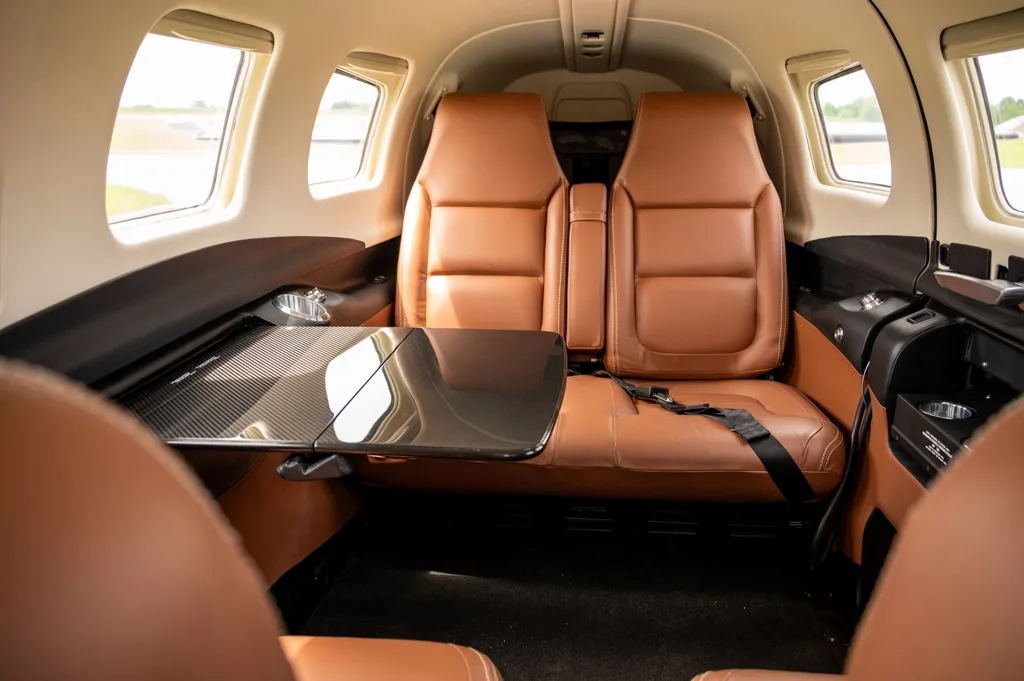The aircraft cabin interior, showing tan leather seats and a fold-out carbon fiber table on the left side.