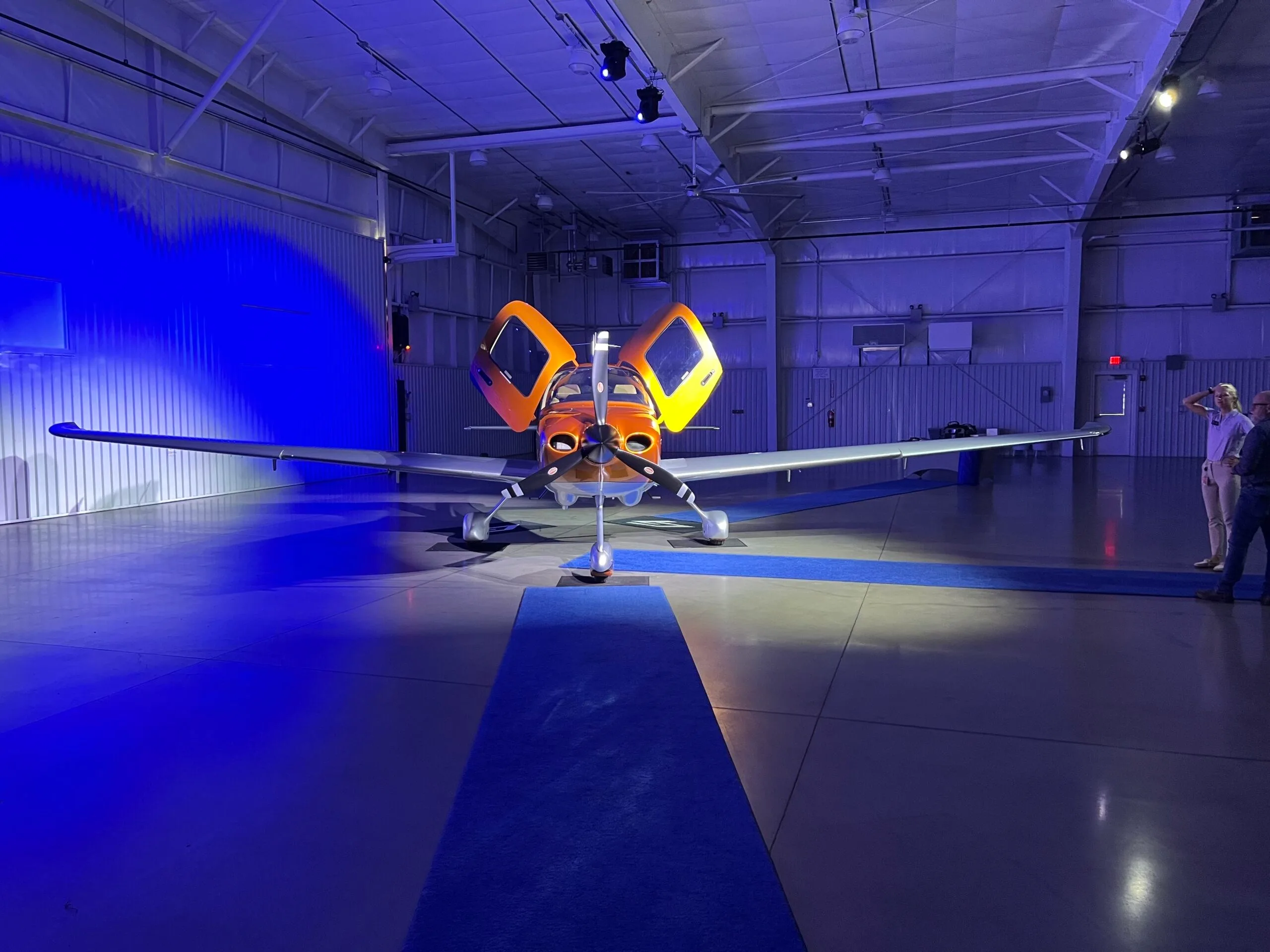 A front view of a Cirrus SR22 aircraft with orange and silver paint, doors open, on a blue carpet in a blue-lit hangar.