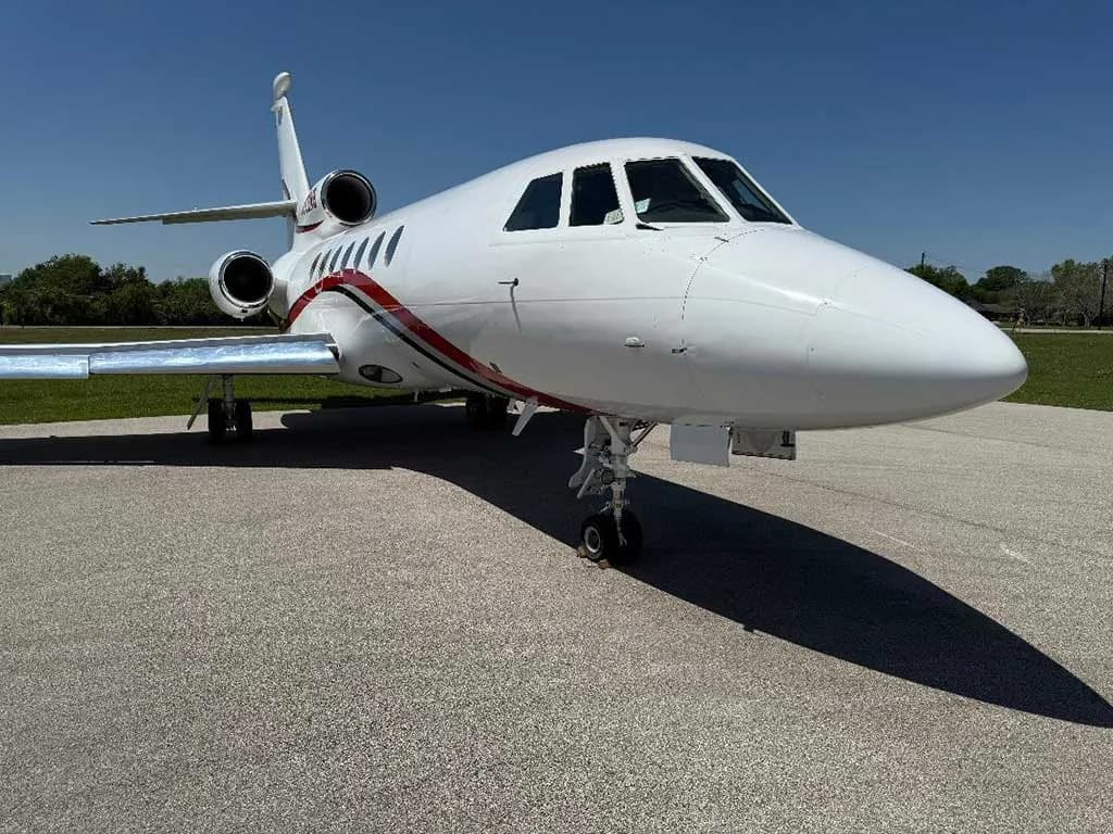 A white Dassault Falcon 900EX business jet with red and black stripes on the tarmac under a clear blue sky, viewed from the front right.