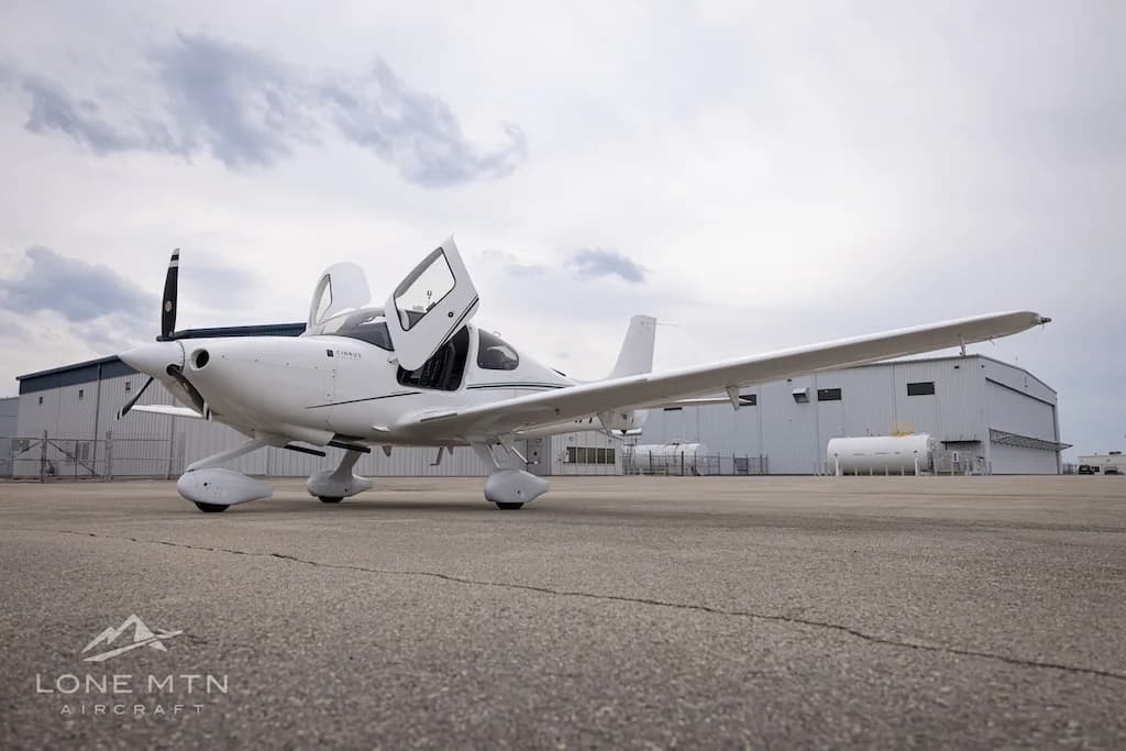 A white Cirrus SR22 aircraft with green accent stripes and open doors, parked on an asphalt ramp.