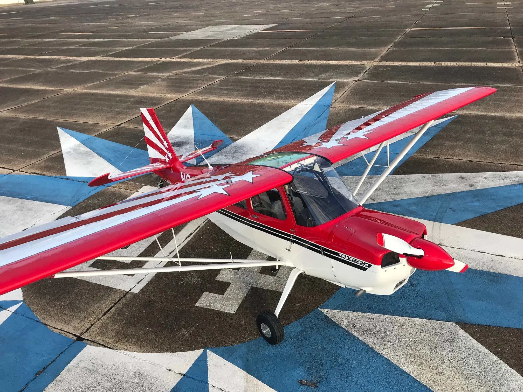 An aerial view of a red and white Super Decathlon aircraft, registration N62WW, parked on a blue and white marked tarmac.