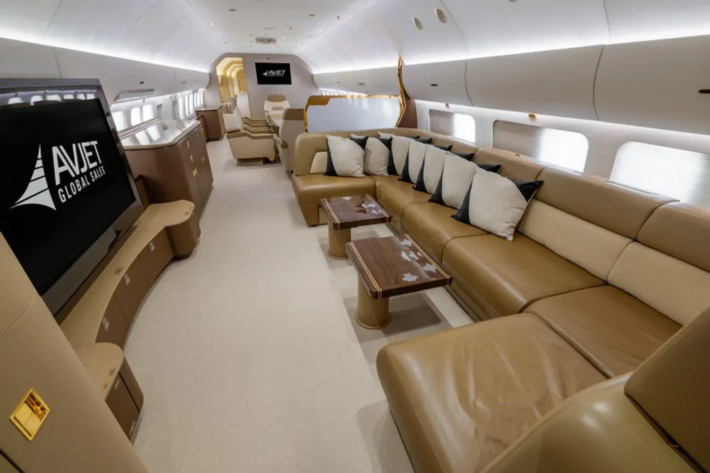 A wide view of a luxurious aircraft cabin featuring a large brown leather sectional sofa with decorative pillows and a large TV screen.