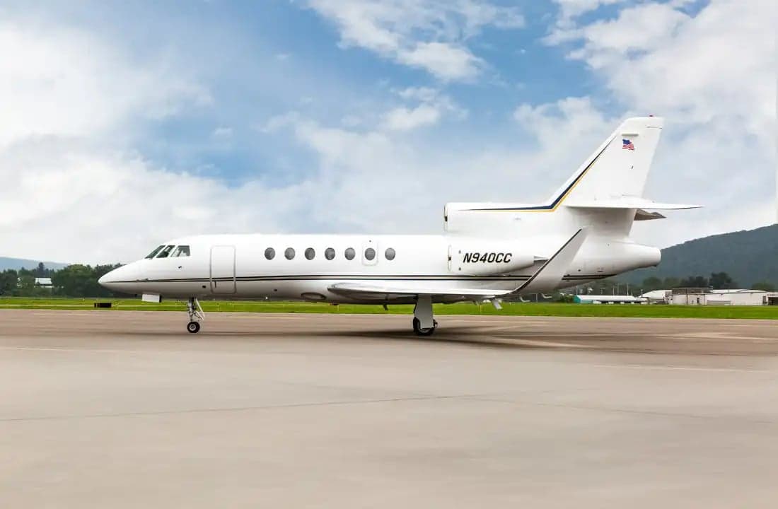 A Falcon jet sits on the tarmac with mountains in the background.