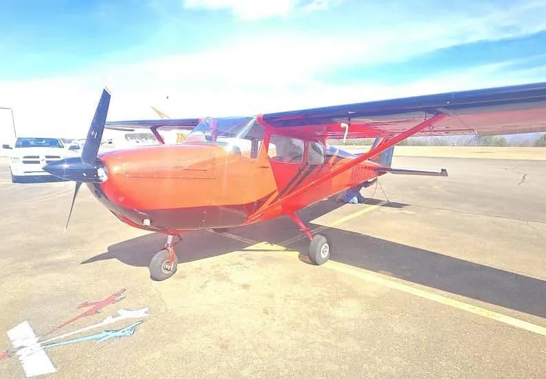 A red and black single-engine propeller aircraft is parked on an airfield with a white truck in the background.