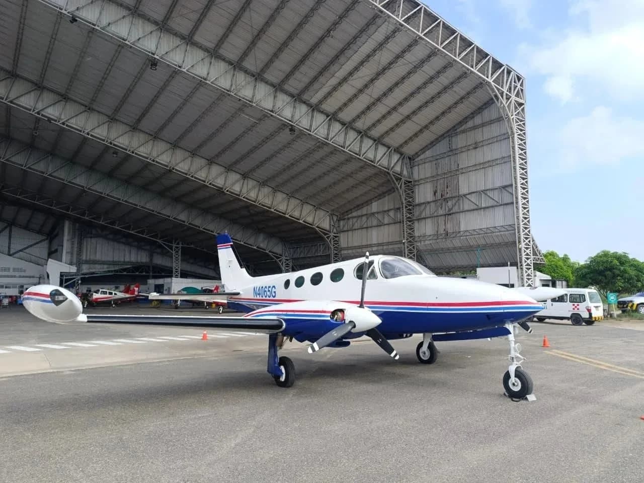 A white and blue Cessna 340 twin-engine aircraft with red stripes parked in front of a large hangar.