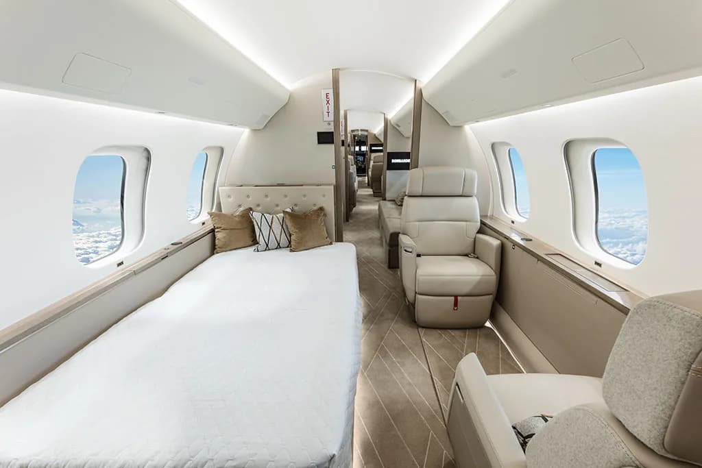 A private jet cabin showing a bed with pillows and a single beige seat, looking towards the rear.