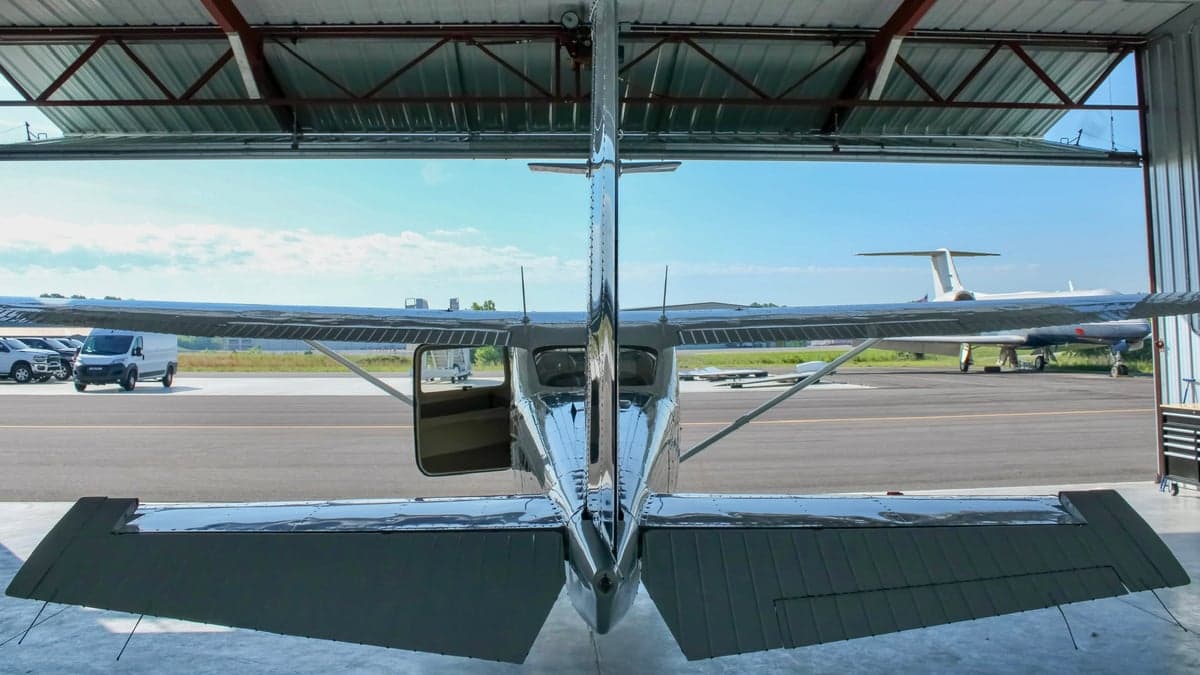 The tail of a white and blue single-engine propeller aircraft, N923DM, inside a hangar, with other aircraft and vehicles visible outside.