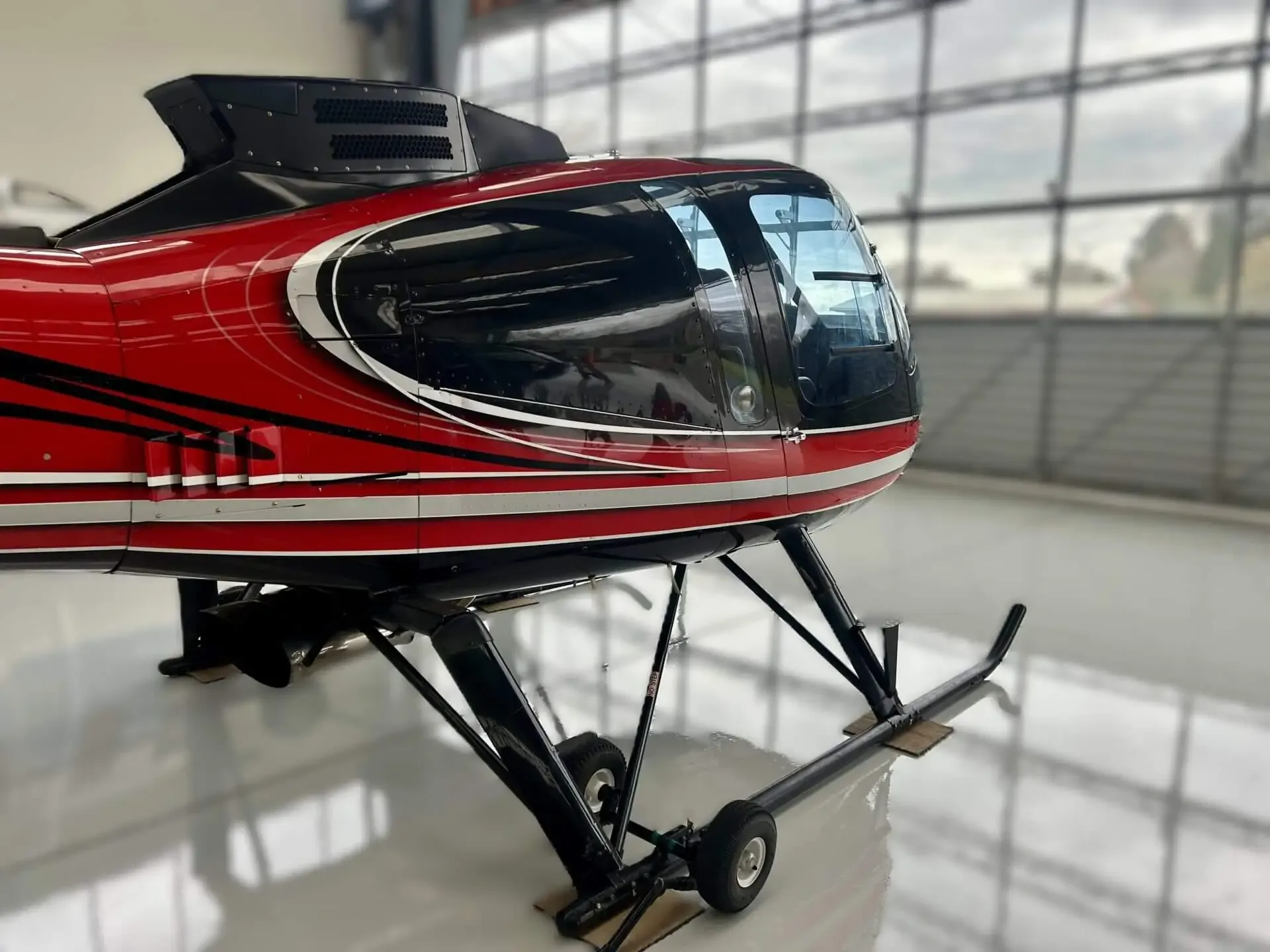 A side view of the front section of the red and black helicopter inside a hangar, showing its cockpit and skids.