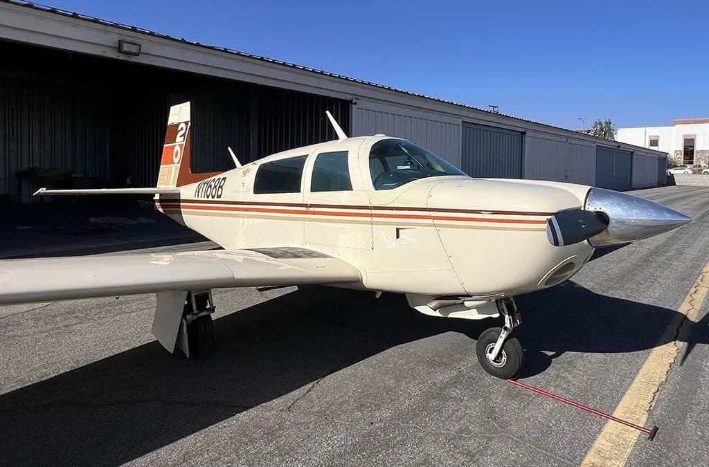 A cream-colored single-engine aircraft with brown and orange stripes, registration N1168B, parked on an asphalt tarmac with hangars in the background.