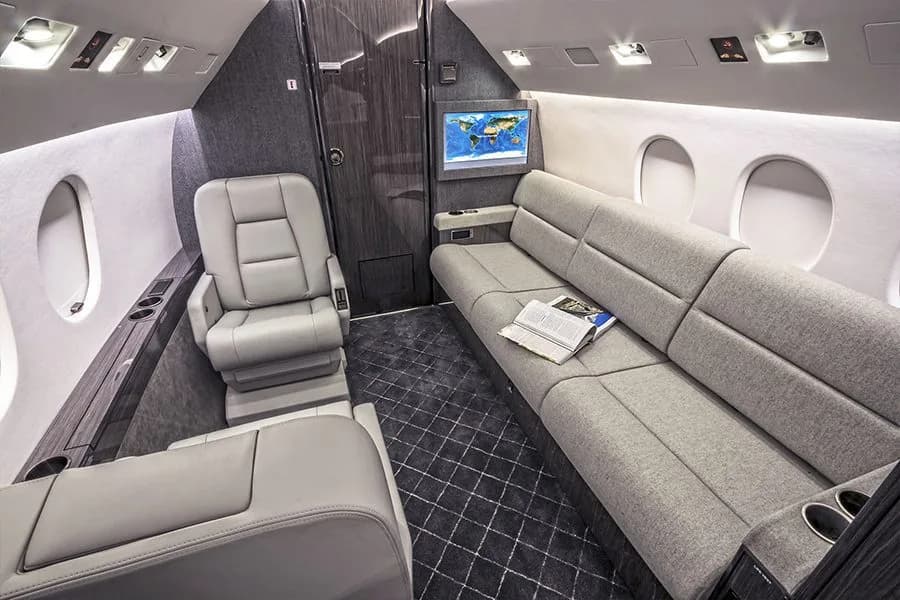 A view of the private jet's cabin featuring a grey sofa, a single seat, and a wall-mounted map display.