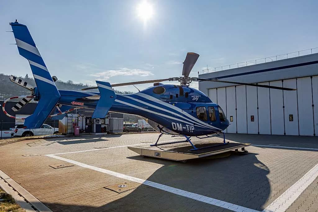 A rear-side view of a blue helicopter with white stripes, registration OM-TIP, on a helipad with the sun in the background.