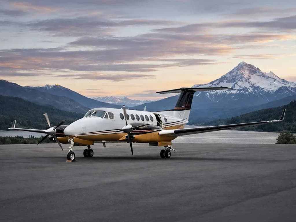 A white and gold Beechcraft King Air turboprop aircraft parked on a tarmac with mountains and a sunset sky in the background.