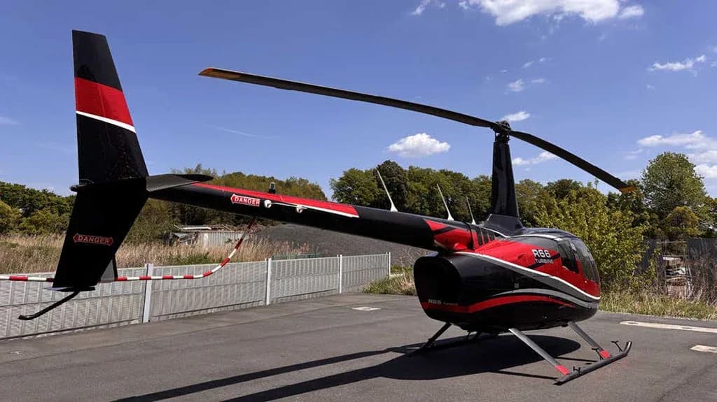 A black and red Robinson R66 Turbine helicopter is viewed from the rear-right, showing its tail rotor and skid landing gear.