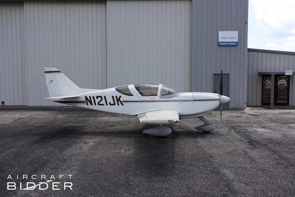 A side view of the white single-engine aircraft (N121JK) with brown stripes parked in front of a hangar.
