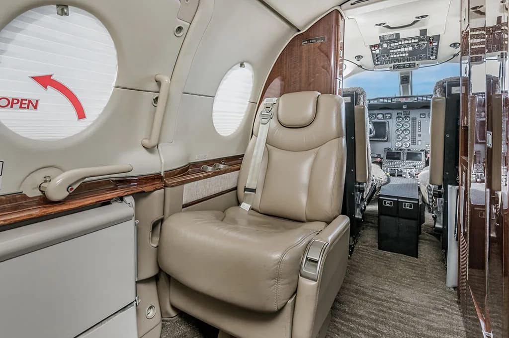A view from the front of the business jet's cabin, showing a beige leather seat and a clear view into the cockpit.