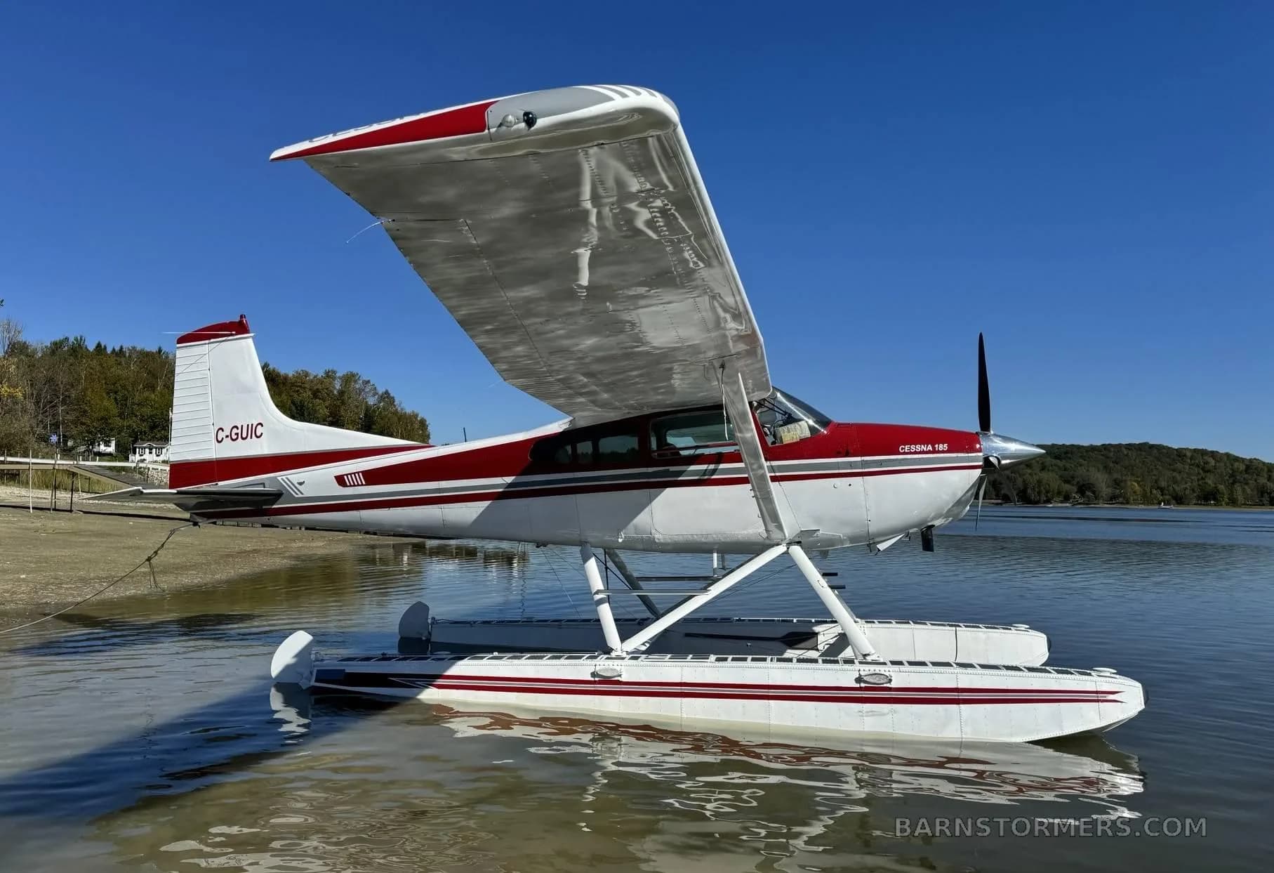 A red and white Cessna 185 seaplane with registration C-GUIC floats on calm water under a clear blue sky.