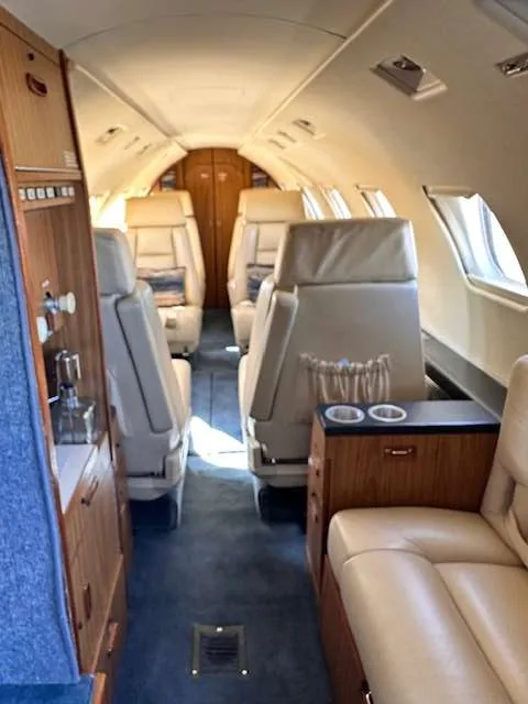 A view down the aircraft's cabin aisle, showing beige leather seats, wood paneling, and blue carpeting.