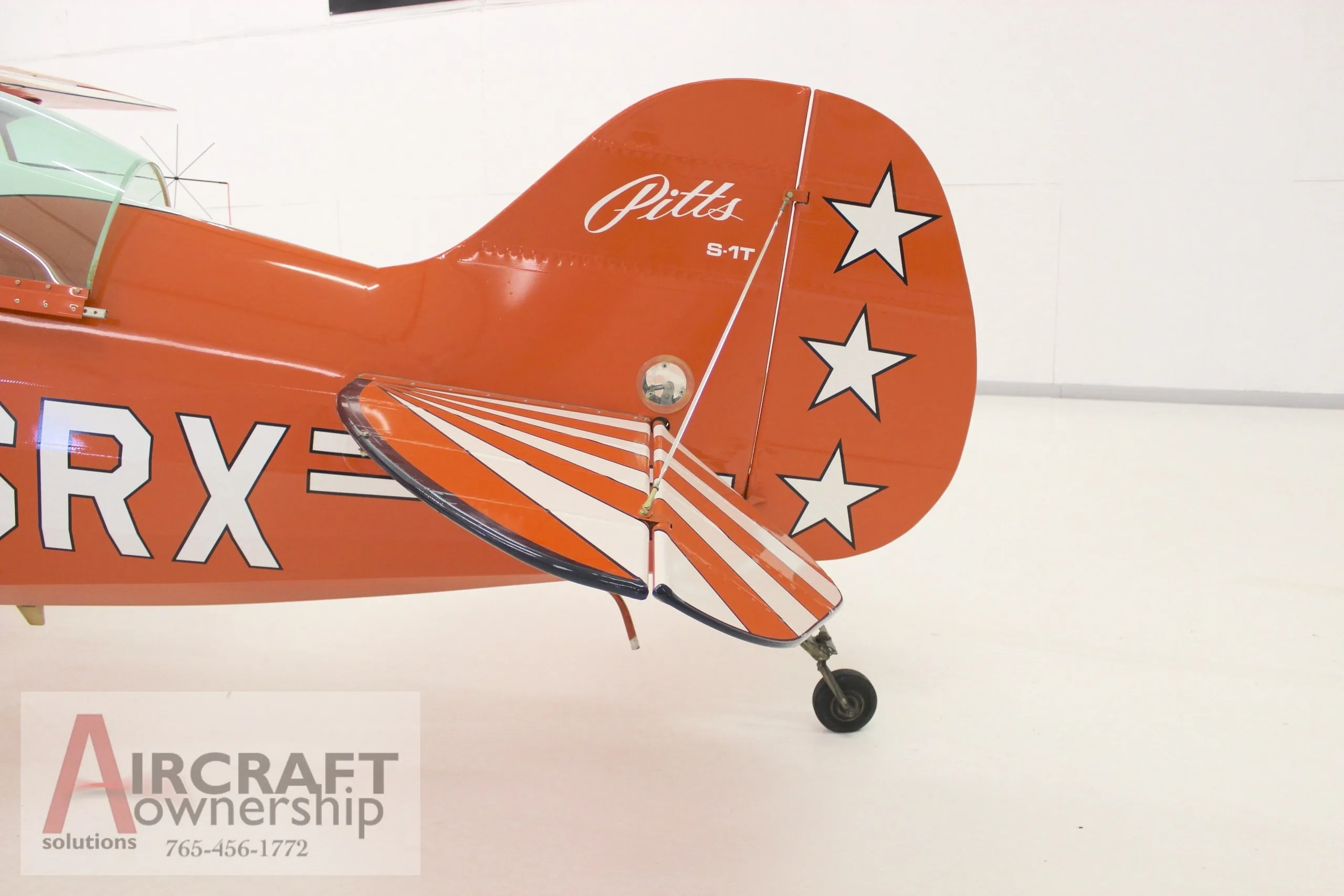 A close-up view of the tail section of an orange Pitts S-1T biplane, showing the tail wheel and white stars on the rudder.
