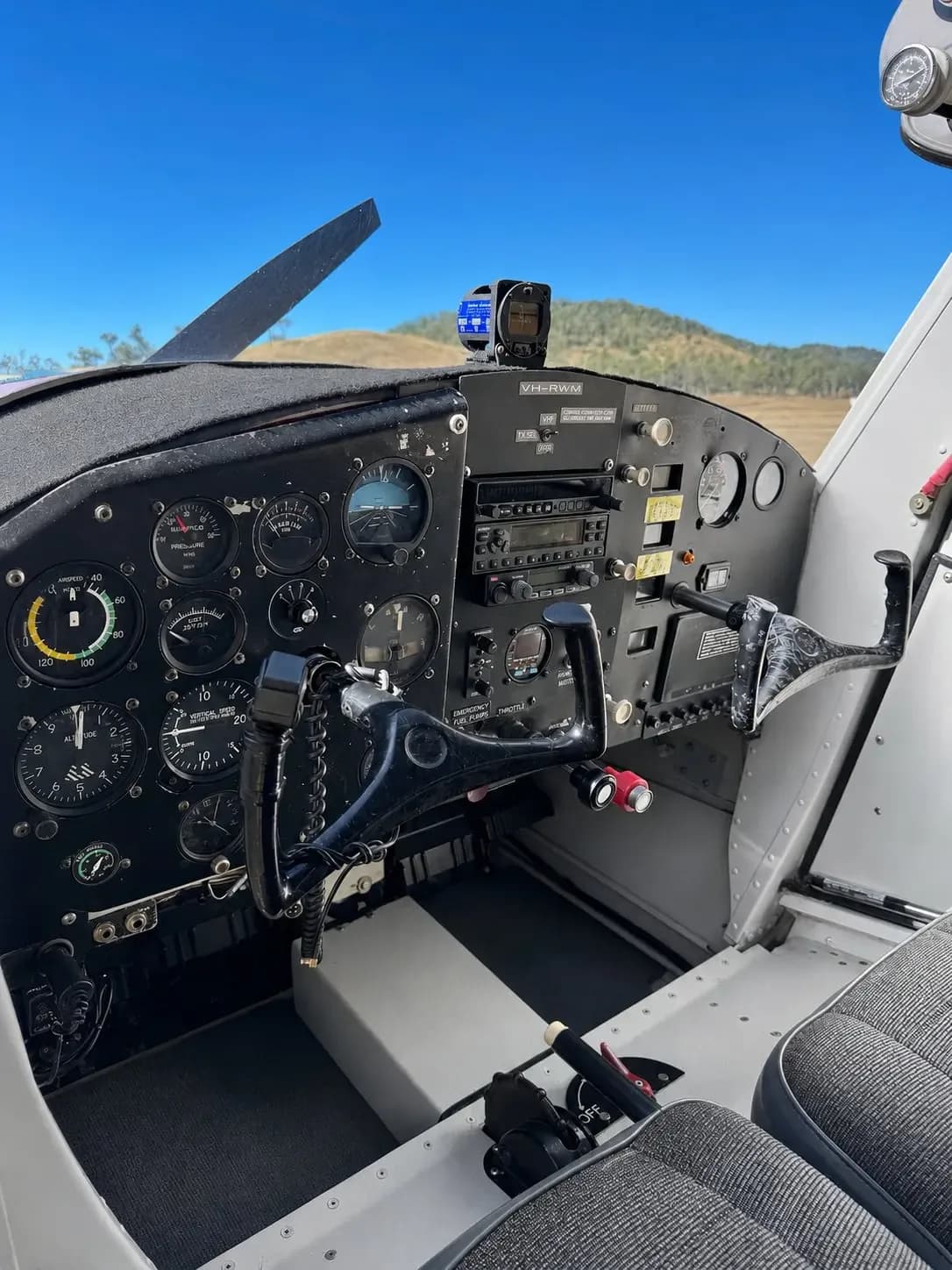 The instrument panel and controls of a small aircraft, featuring analog gauges and a yoke, with a view of hills outside.