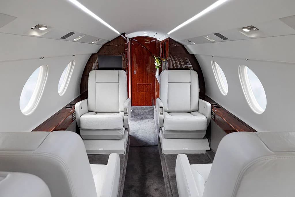 The luxurious interior cabin of a business jet, featuring light gray leather seats and dark wood accents.