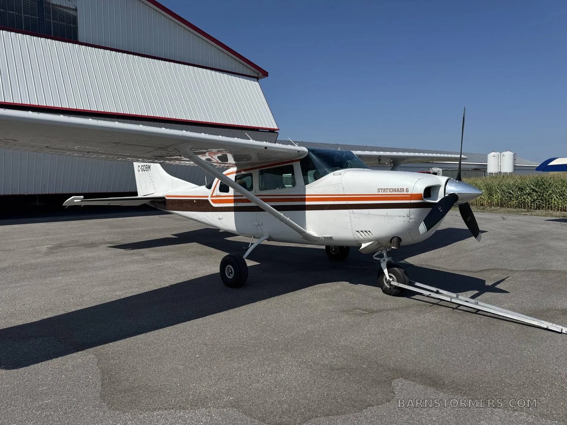 A white Cessna Stationair 6 with brown and orange stripes parked on an asphalt ramp next to a white hangar.