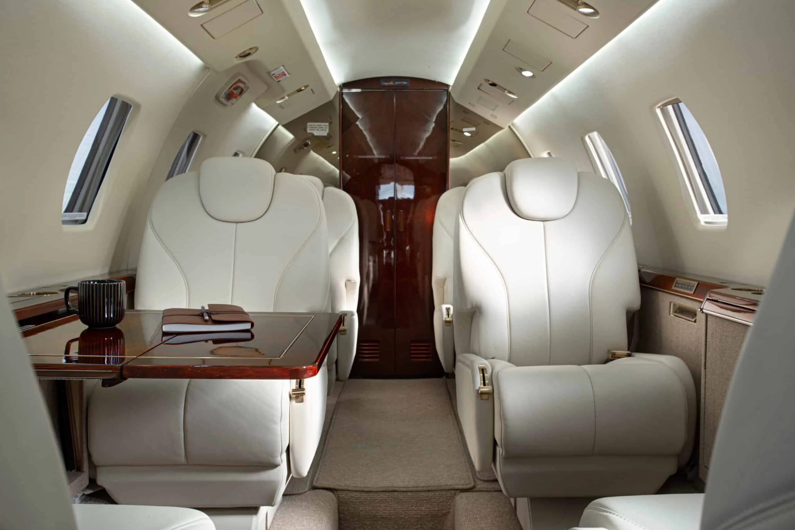 A view of the aircraft cabin with two cream leather seats facing each other, a polished wooden table, and overhead lighting.