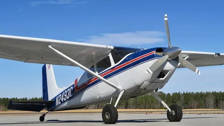 A white and blue Cessna 180 Skywagon with red stripes, registration N2493C, parked on an airfield under a clear blue sky.