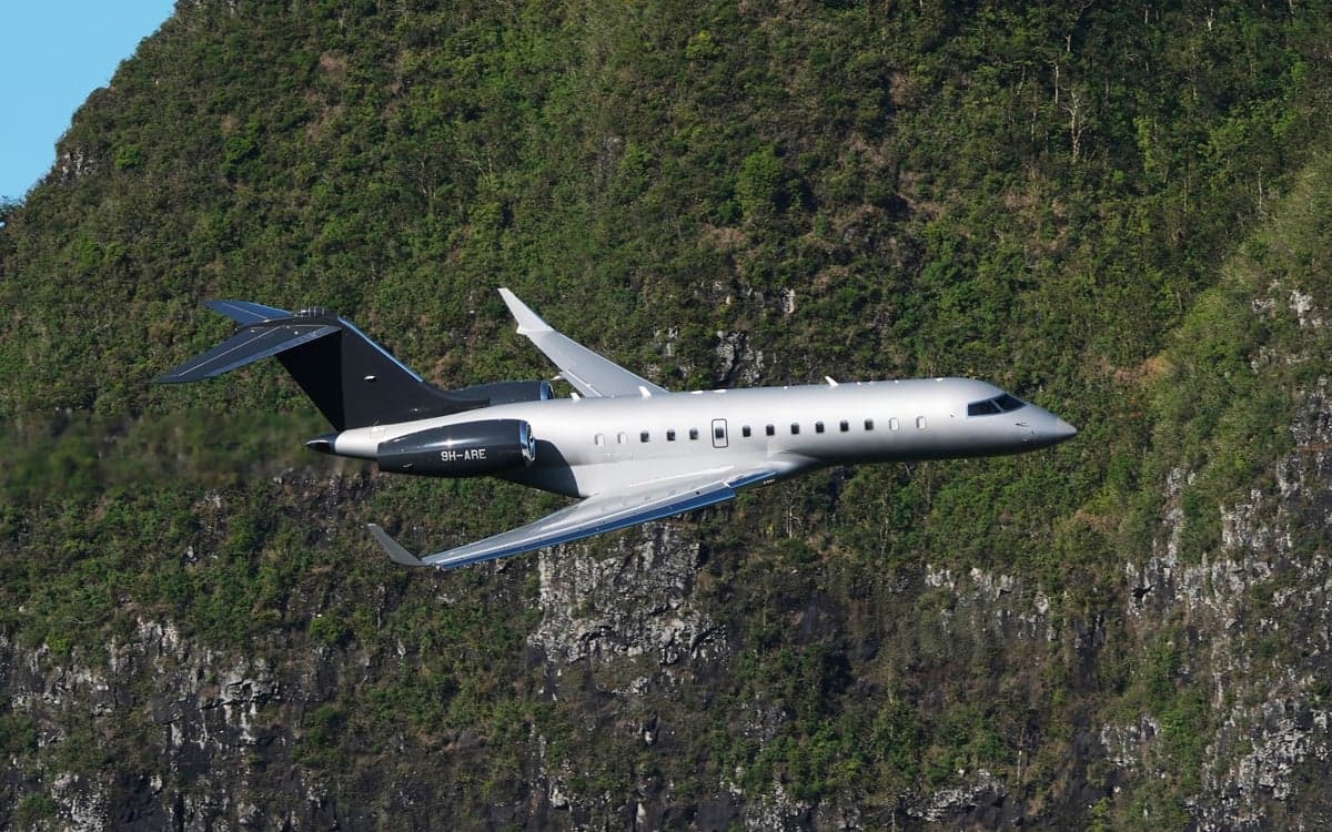 The Bombardier Global 5000 aircraft 9H-ARE is shown in flight against a backdrop of lush green mountains.