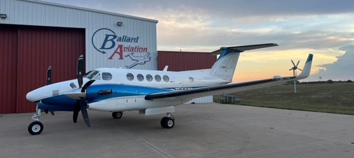 A King Air turboprop aircraft is parked outside a hangar with a blue and white paint scheme.