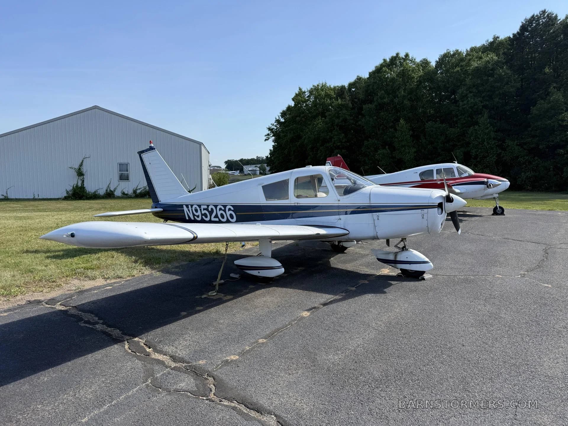 A white single-engine propeller aircraft (N95266) with blue stripes, viewed from the side-rear, parked on an airport apron with trees and a hangar.