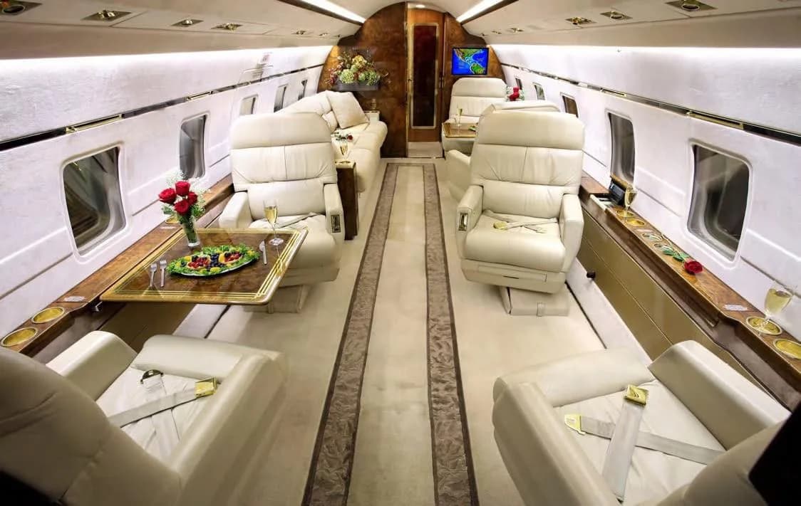 A luxurious aircraft cabin with cream leather seats, a dining table set with fruit, and wood accents.