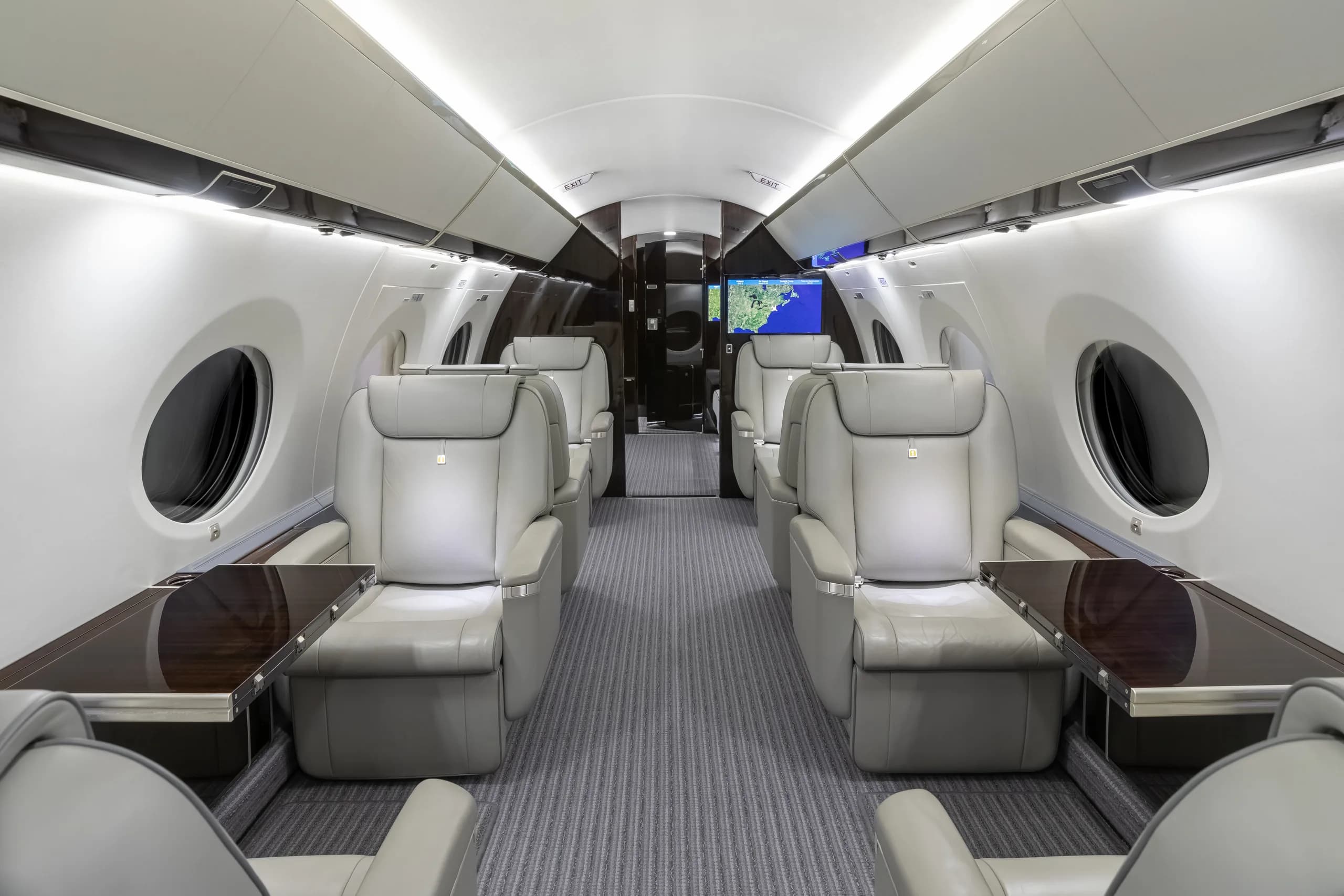 A wide view of the private jet's main cabin, featuring rows of light grey leather seats and dark wood tables.