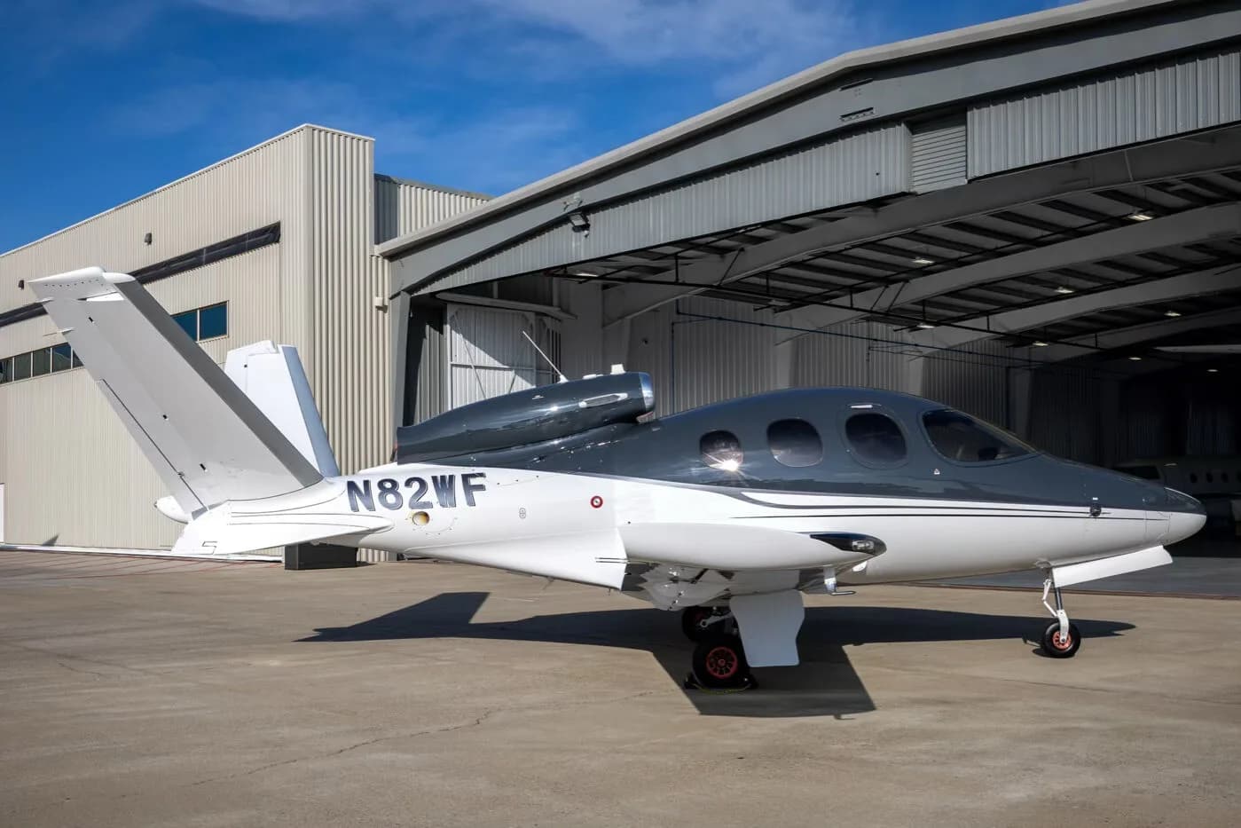 A three-quarter rear view of a white and dark grey Cirrus Vision Jet (N82WF) parked outside a hangar.