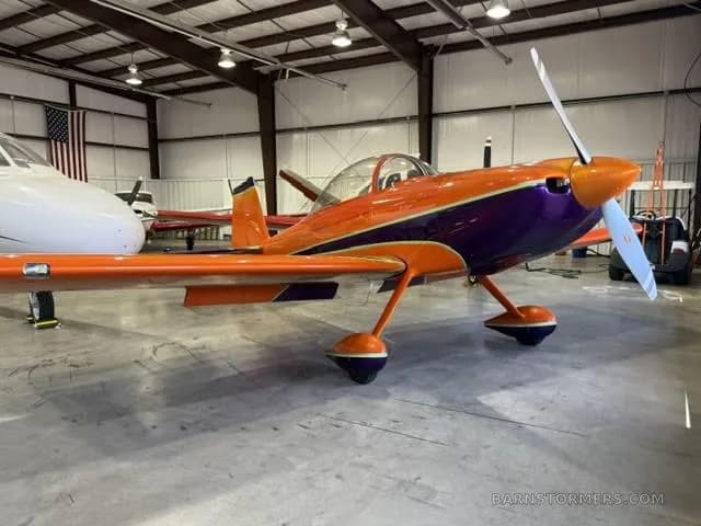 A three-quarter front view of an orange and purple single-engine aerobatic aircraft parked in a hangar with another plane.