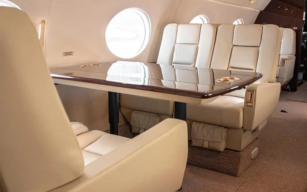 A view of an aircraft cabin with two sets of facing beige leather seats and a large polished wooden table between them.