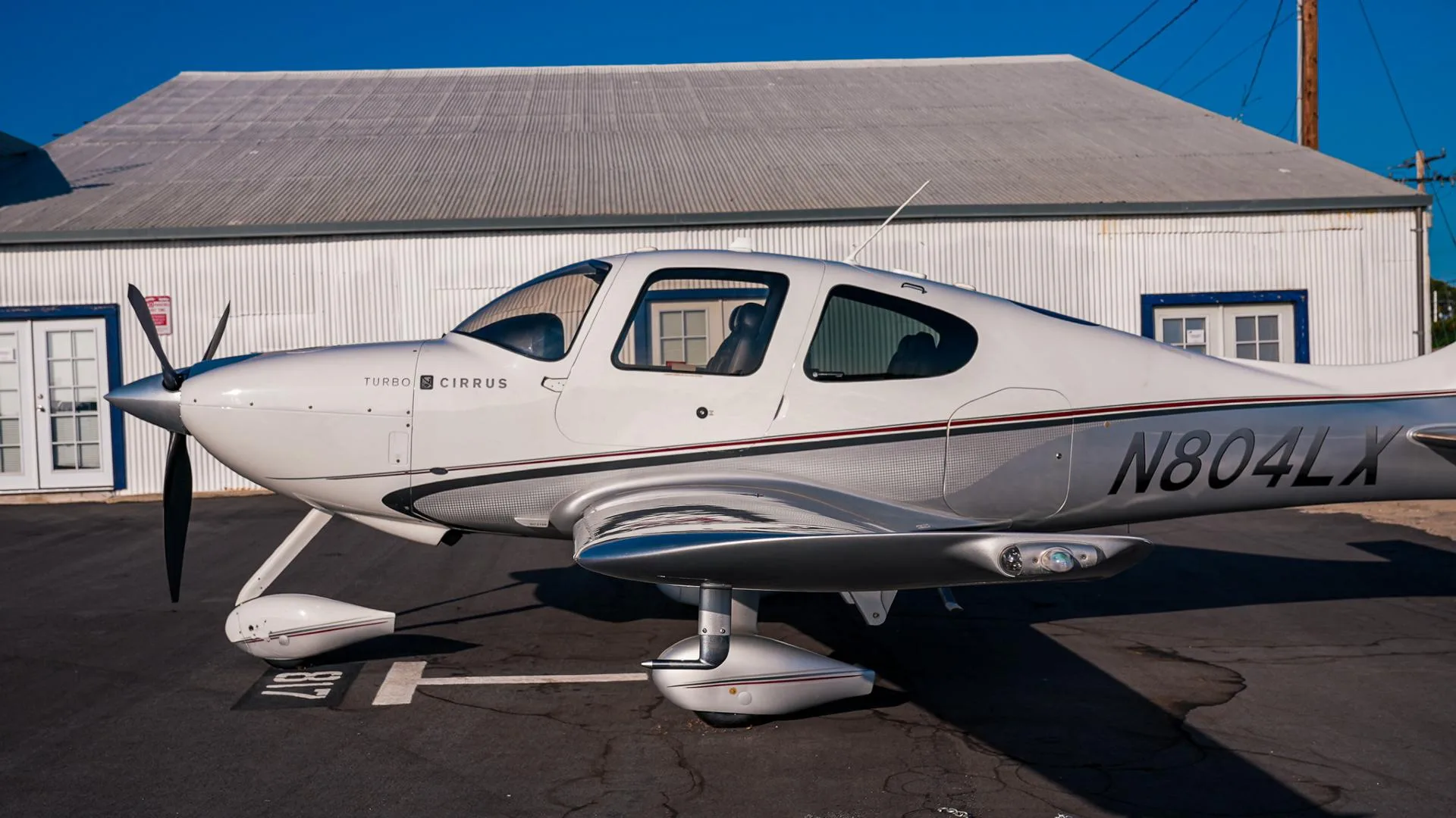 A side view of the white and silver Cirrus SR22T aircraft N804LX with a hangar in the background.