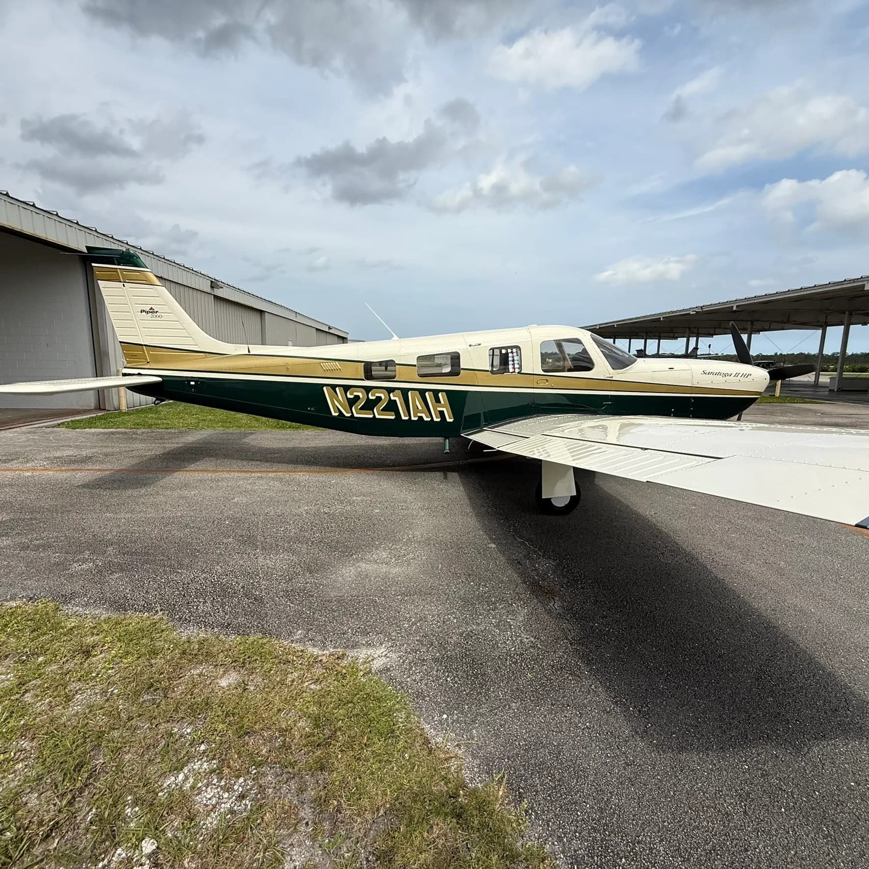 A Piper Saratoga II HP, N221AH, with a white, gold, and dark green paint scheme, parked on an airport tarmac with hangars in the background.