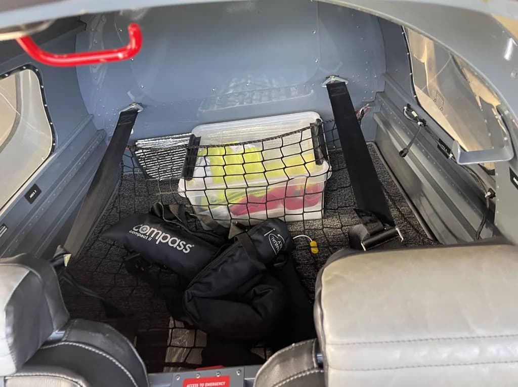 The baggage compartment behind the seats of a Sonaca 200 aircraft, secured with a net and containing a plastic bin and bag.