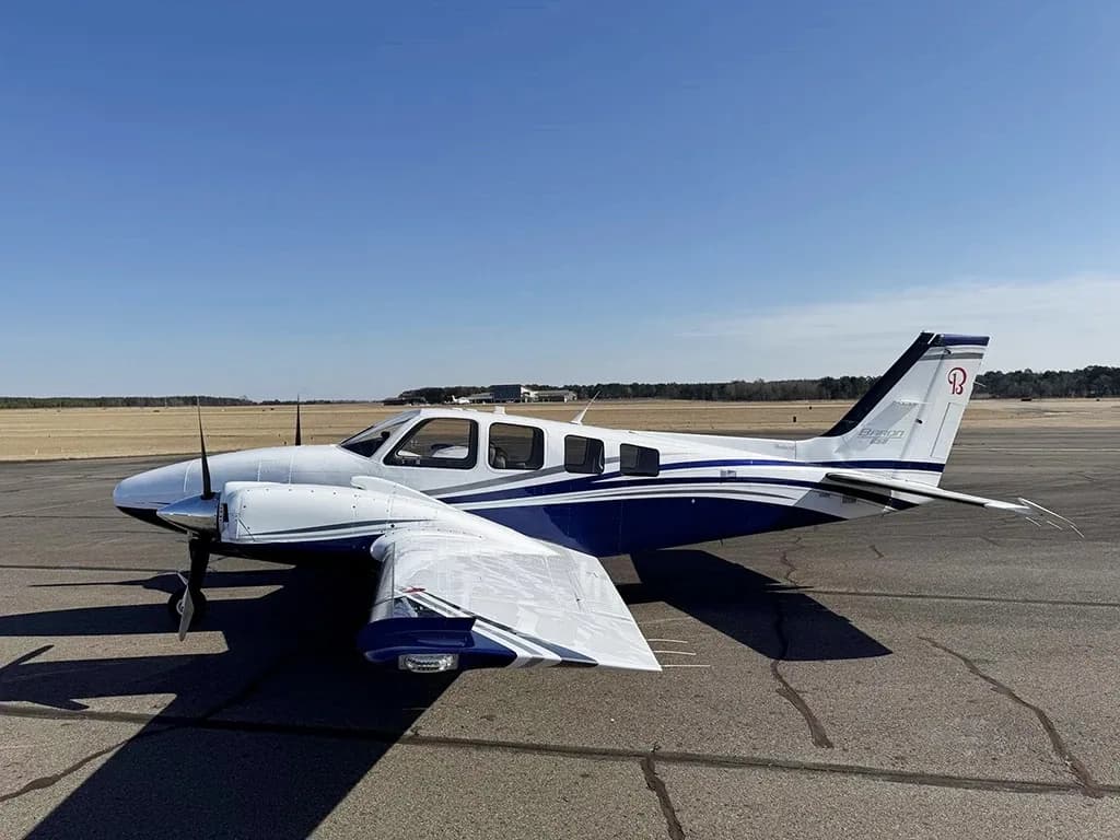 A white and blue Beechcraft Baron 55 twin-engine aircraft is parked on an airport tarmac under a clear sky.