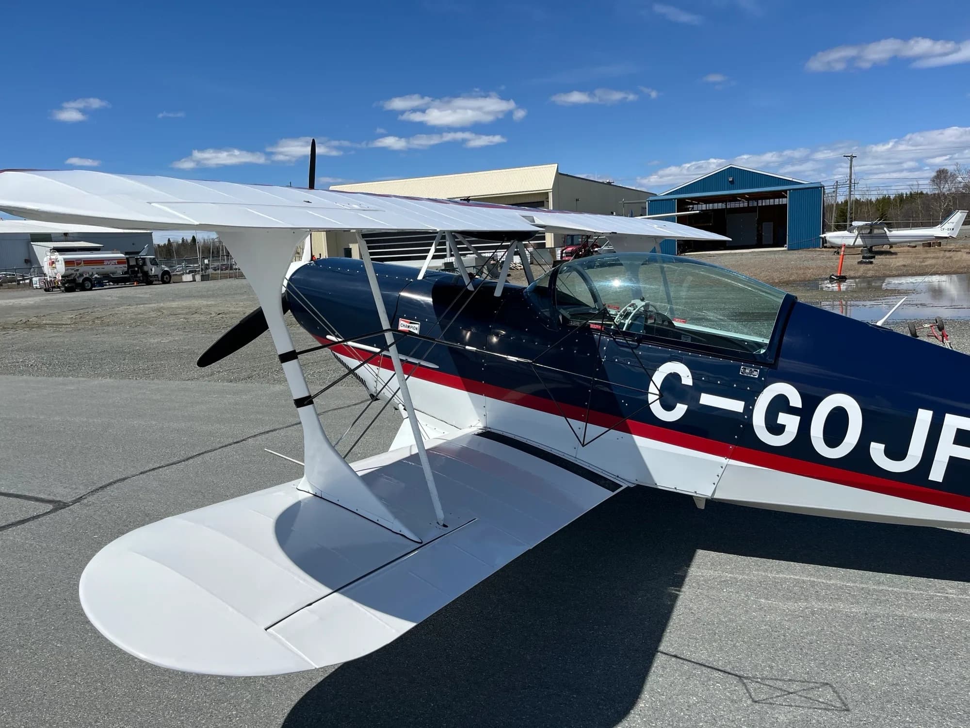 A close-up side view of the dark blue and white Pitts biplane C-GOJF on an airfield with hangars in the background.