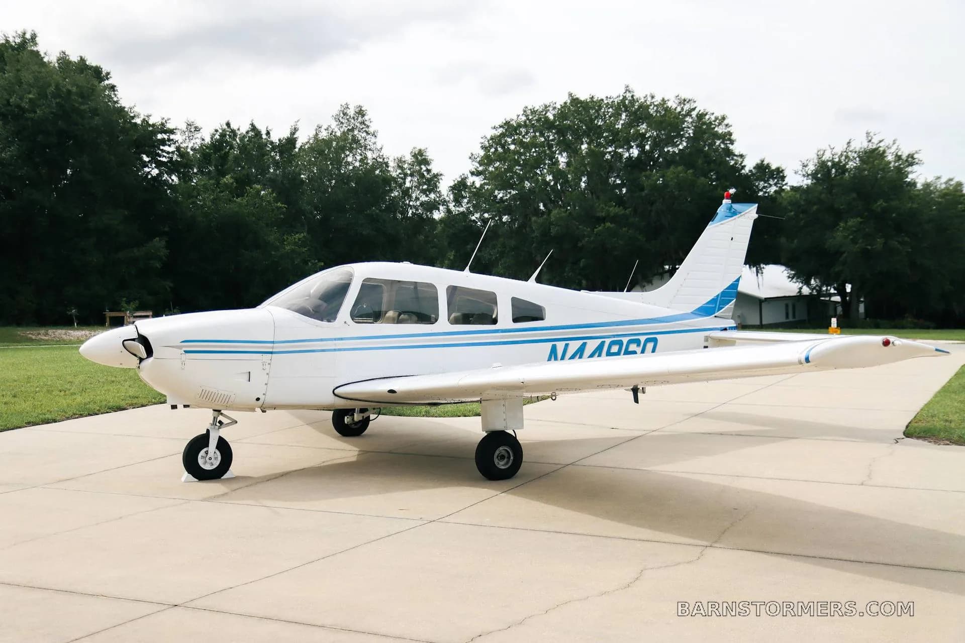 A white Piper PA-28 series aircraft with blue stripes, viewed from the front-right, parked on a concrete pad.