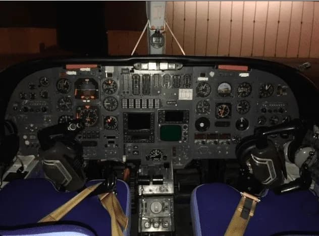 A detailed view of an aircraft cockpit with numerous analog gauges, control yokes, and blue seats.