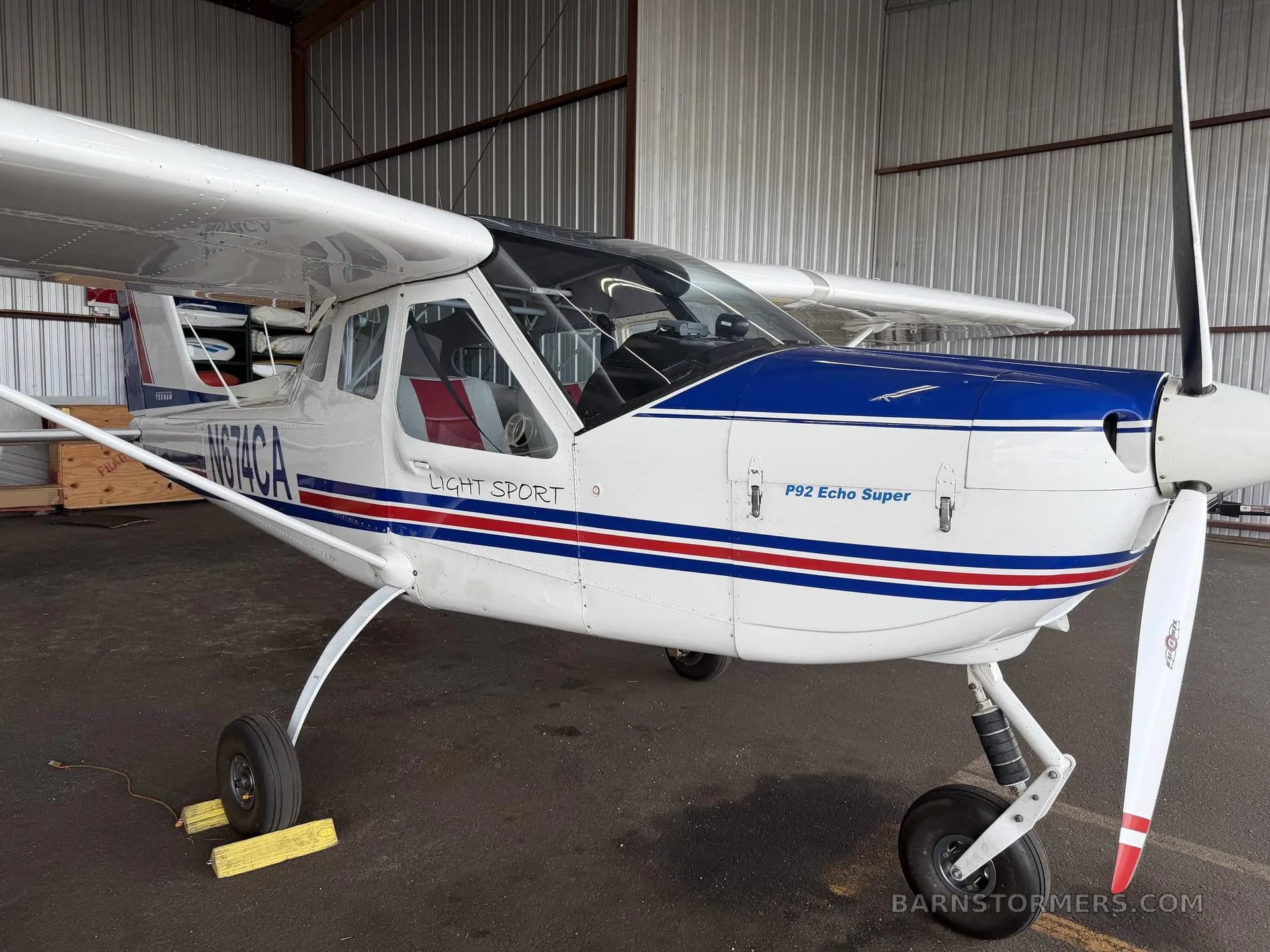 A white Tecnam P92 Echo Super light sport aircraft with blue and red stripes, parked in a hangar.