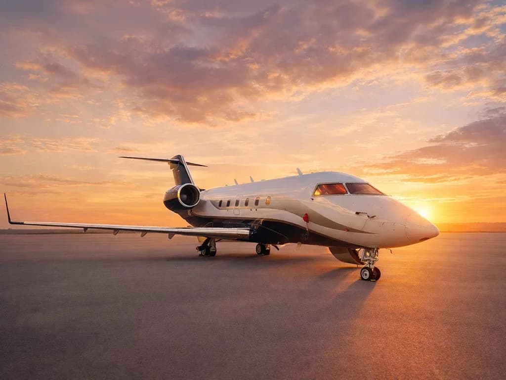 A white and dark blue private jet is parked on an airfield at sunset, with warm orange and pink clouds in the sky.