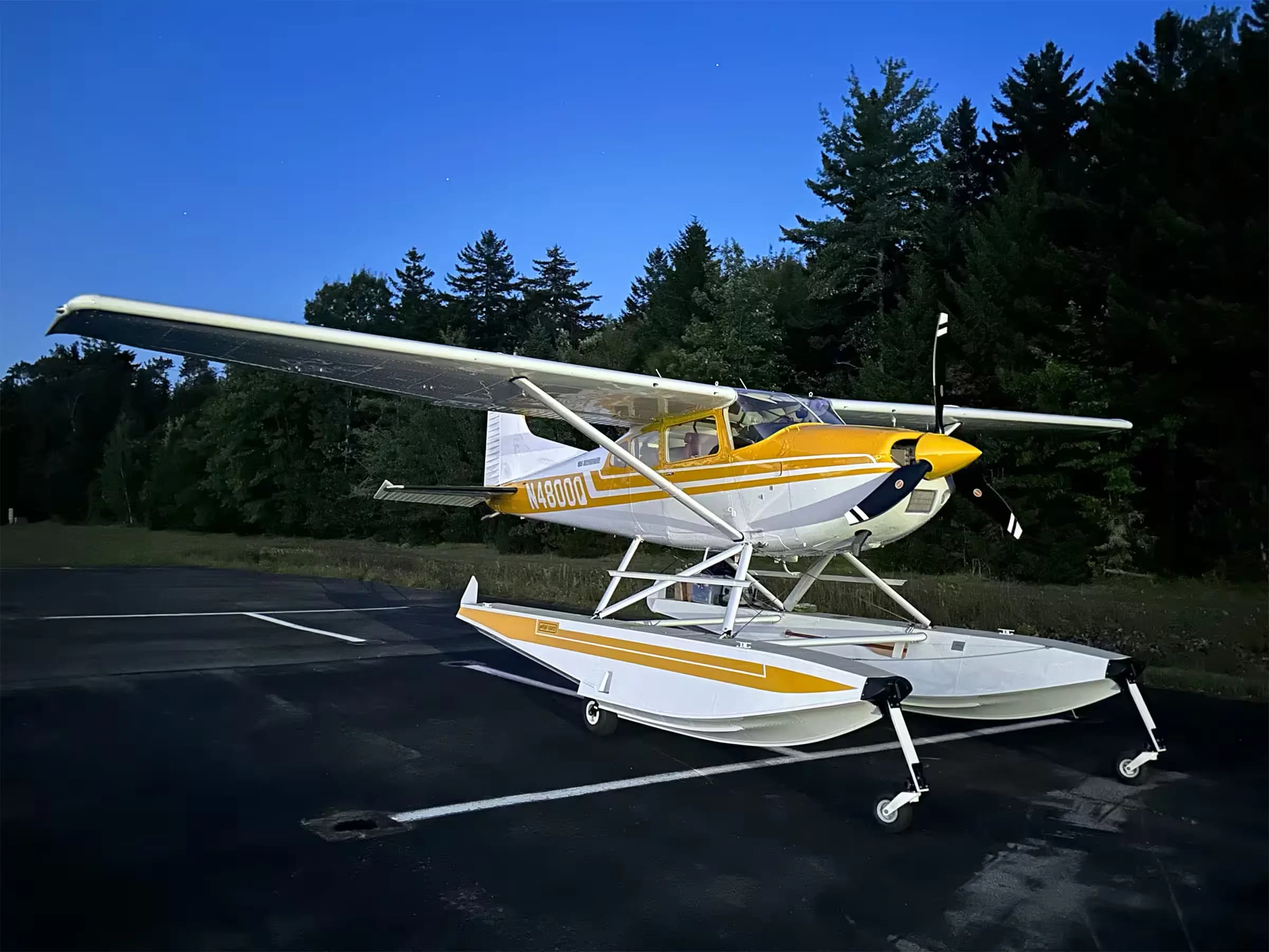 A yellow and white Cessna 185 floatplane, N4800Q, parked on an asphalt ramp at dusk with trees in the background.