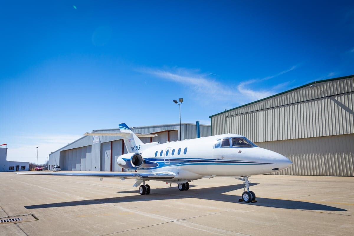 A white and blue jet sits on the tarmac in front of a hangar on a sunny day.